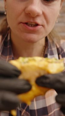 Dikey video. Siyah Eldivenli Kadın Peynirli Burger 'i Isırır, Yanında Patates Kızartması Yiyen Kadın.