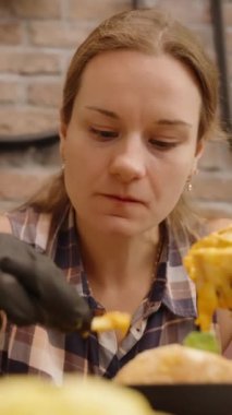 Dikey video. Siyah Eldivenli Kadın Fransız kızartması yiyor, Samimi bir kafede peynirli burgerden bir ısırık alıyor..