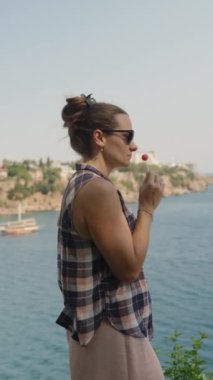 Dikey video. Kadın lolipopun tadını çıkarıyor, güneşli bir günde sahnenin manzarasını seyrediyor..