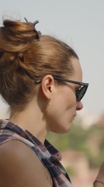 Dikey video. Güneş Gözlüklü Kadın Lolipopun Tadını Çıkartıyor, Güzel Manzaralı Rıhtımda Dinleniyor.