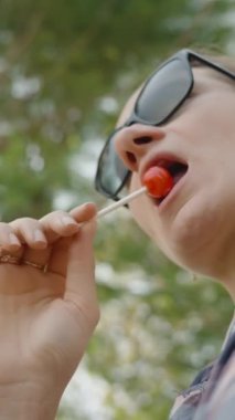 Dikey video. Güneş Gözlüklü Kadın Lolipopun Tadını Çıkarıyor, Açık Hava Sahnesini Çekiyor.