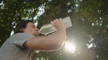 Adam Şişe 'den su içiyor, Parlak Güneş Işığıyla Ağaçların Altında Tazeleyici Molasının tadını çıkarıyor.