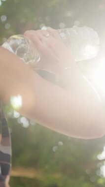 Dikey video. Kadın, sıcak bir yaz gününde ağaçların arasından süzülen parlak güneş ışığıyla kendini sulandıran şişeden su içer..