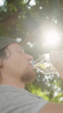 Dikey video. Adam Şişe 'den Suyu İçti, Gün Işığı Parkı' nda Ağaçların arasından Parlayan Güneş Işığı ile kendini tazeledi.