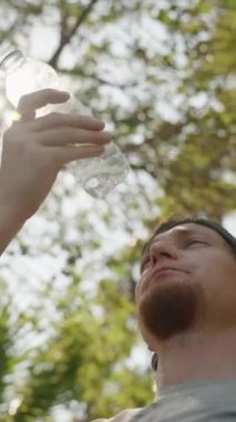 Dikey video. İnsan Şişe 'den su içer, Güneş Işığı ve Ağaçların Altındaki Serinletici İçkinin tadını çıkarır.