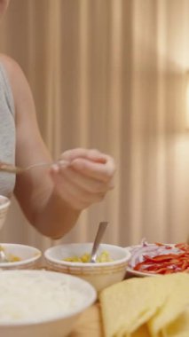 Dikey video. Kadın rahat bir evde taco hazırlıyor, taco kabuğuna domates sosu ekliyor..