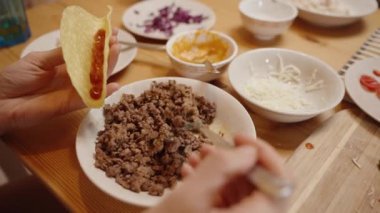 Bir Kişi Taco Kabuğuna Kıyma Ekler, Etrafı Peynir, Salsa ve Lezzetli Yemek İçin Taze Malzemelerle çevrili.