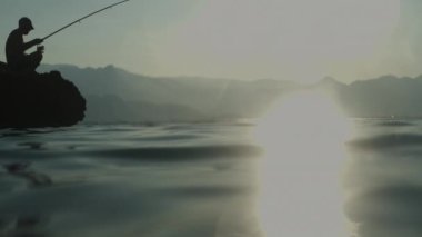Batan Güneş 'e Karşı Yüzen Bir Balıkçı Silueti Halatta Yakalandı. Dağların arka planında, denizde bir kayanın üzerinde oturmak. Yavaş çekim.
