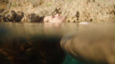 Rocky Shores 'ın yanında Yüzen Bir Kadının Profili, Huzurlu Rahatlamayla Kapalı Gözleriyle Suyun İçinden Güneş Işığı Süzülürken. Yavaş çekim.