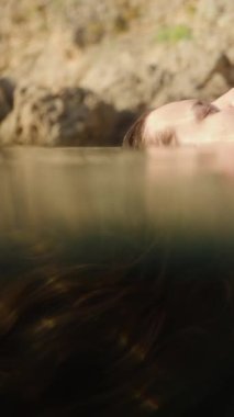 Dikey video. Rocky Shores 'ın yanında Yüzen Bir Kadının Profili, Huzurlu Rahatlamayla Kapalı Gözleriyle Suyun İçinden Güneş Işığı Süzülürken. Yavaş çekim.