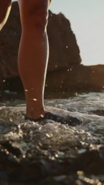 Dikey video. Su Ayakkabıları İçinde Kayalık Arazi, Sakin Deniz ve Altın Işık Altında Kayalık Kayalıklar Üzerinde Tedbirli Yürüyen Bir Kişi. Yavaş çekim.