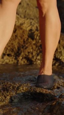 Dikey video. Su Ayakkabıları 'nın Yavaş Hareketi Engebeli Kayalıklarda Yürüyor, Güneş Işığı Islak Yüzeyde Altın Renk Oluyordu. Yavaş çekim.