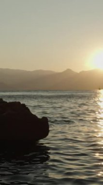 Dikey video. Güneş Dağların Ardında Batarken Rocky Shore 'da durup Deniz' e Altın Parıltı saçan Bir Kadın Göz kamaştırıcı Günbatımını Telefonuyla Yakalıyor. Yavaş çekim.