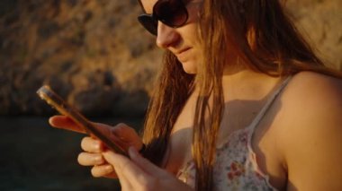 Güneş Gözlüklü Bir Kadın Telefonuna Odaklanıyor, Kayalık Bir Arkaplanda Duruyor, Gün batımının Ilık Işığı Yoğunlaştırılmış İfadesini Vurguluyor. Yavaş çekim.