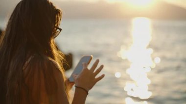 Bir Kadın Telefonunu Deniz kenarında Günbatımında Kullanıyor, Sudan yansıyan Altın Işık, Huzurlu ve Huzurlu bir atmosfer Oluşturuyor.