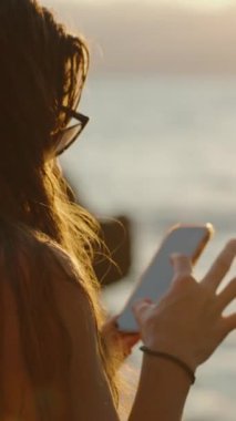 Dikey video. Bir Kadın Telefonunu Deniz kenarında Günbatımında Kullanıyor, Sudan yansıyan Altın Işık, Huzurlu ve Huzurlu bir atmosfer Oluşturuyor.