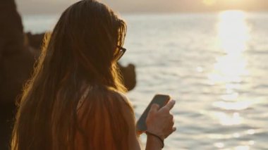 Sahil şeridinde Duran Bir Kadın Telefonunu Kullanıyor, Gün batımı Sudan Yansıyarak Sıcak ve Huzurlu Bir Sahne Yaratıyor.