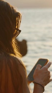 Dikey video. Sahil şeridinde Duran Bir Kadın Telefonunu Kullanıyor, Gün batımı Sudan Yansıyarak Sıcak ve Huzurlu Bir Sahne Yaratıyor.