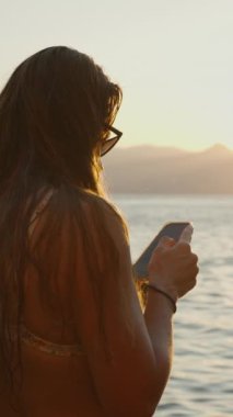 Dikey video. Güneş batarken Denizde Duran Bir Kadın, Telefonunu kullanarak, Sıcak Altın Işıkla Sudan Yansıyan ve Çevresindeki Kayaları Yansıtan.