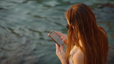 Bir Kadın Telefonuna Suyun Yanında Bakar, Islak Saçları ve Güneş Gözlükleri Doğal Işıkla Vurgulanır, Huzurlu Bir Sahil Sahnesi Oluşturur.