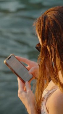 Dikey video. Bir Kadın Telefonuna Suyun Yanında Bakar, Islak Saçları ve Güneş Gözlükleri Doğal Işıkla Vurgulanır, Huzurlu Bir Sahil Sahnesi Oluşturur.