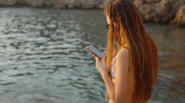 Suyun Kıyısında Duran Bir Kadın Telefonunu, Sakin Deniz 'i ve Huzurlu Bir Arkaplan Oluşturan Rocky Shoreline Bakıyor.