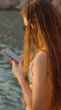 Dikey video. Suyun Kıyısında Duran Bir Kadın Telefonunu, Sakin Deniz 'i ve Huzurlu Bir Arkaplan Oluşturan Rocky Shoreline Bakıyor.