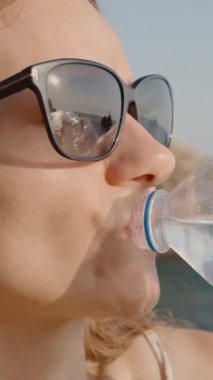 Dikey video. Güneş Gözlüklü Kadın Plastik Şişeden Su İçiyor, Deniz ve Uçurumlar Lenslerine Yansıtılıyor, Açık Hava Gününün Tadını Çıkartıyor.