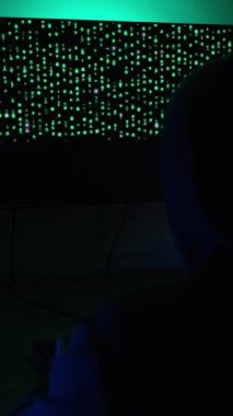 Dikey video. Kapüşonlu Tişörtlü Adam Karanlık Oda 'da oturuyor, Yeşil Matris benzeri Kod gösteren bir monitörle yüzleşiyor, Hacker Vibe yaratıyor..