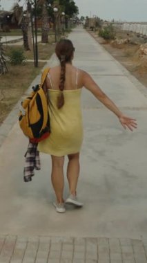 Dikey video. Sarı havluya sarılı, sırt çantalı ve yüzdükten sonra tişörtlü bir kadın sarılı bir şekilde taşlı bir yolda yürüyor..