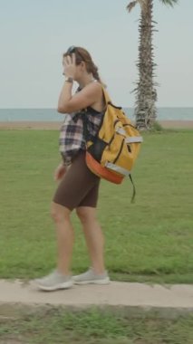 Dikey video. Sahile yakın çimenli bir yolda yürüyen, sırt çantası takan palmiye ağaçları ve arka planda okyanus olan bir kadın..