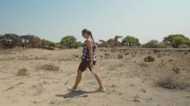 Bir kadın çorak arazide yürüyor, kumlu arazide, Bir şapka ve şişe tutuyor, Uzakta dağılmış ağaçlarla.