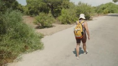 Yeşil çalılar ve ağaçlarla kaplı, Asfalt bir yolda yürüyen Şapka ve Sırt Çantası Giyen Bir Kadın.
