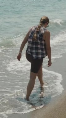 Dikey video. Bir kadın kıyı şeridi boyunca yalınayak yürüyor, Islak Kumda yürürken ayaklarının üzerinde nazikçe dalgalar süzülüyor.