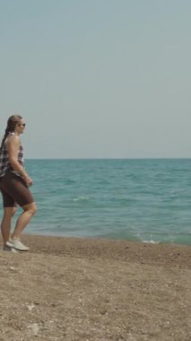 Dikey video. Bir kadın kumsalda yürüyor, okyanus manzarasının tadını çıkarıyor, uzak sularda görünen teknelerle..