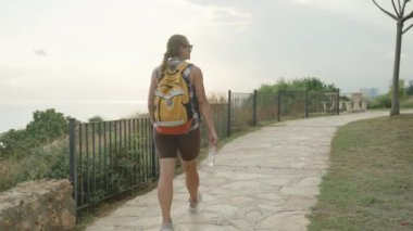 Okyanus manzaralı taştan bir yolda yürüyen, elinde bir su şişesi tutan ve öğleden sonra güneşinin tadını çıkaran bir kadın..