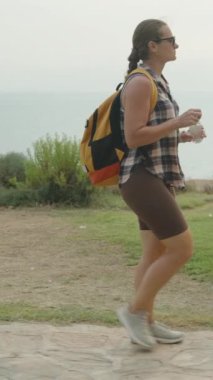 Dikey video. Bir Kadın Taş Yolda Yürür, Yanında Okyanus, Huzurlu Gezintide Bir Su Şişesi Taşır.