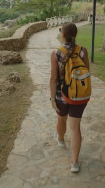 Dikey video. Deniz kıyısındaki taş bir yolda yürüyen bir kadın, elinde bir su şişesiyle huzurlu bir yürüyüşün tadını çıkarıyor..