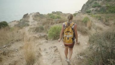 Kıyıya yakın engebeli bir yolda yürüyen bir kadın elinde bir su şişesi tutuyor. Arka planda Uzak Tepeler ile Kuru Bitkiler Arasında Yol Rüzgarları.