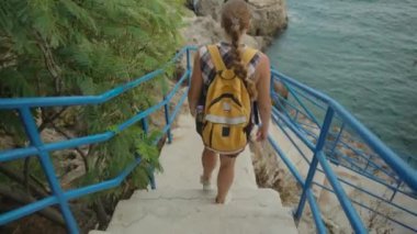 Bir kadın mavi raylı taş merdivenlerden suya doğru iniyor. Yol, Rocky Kıyı Şeridi ve Sakin Deniz 'in Manzarasını Sunar.