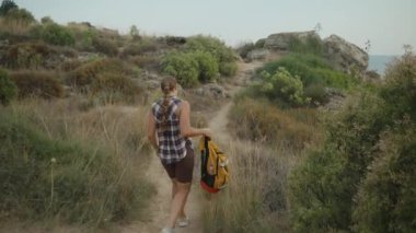 Kayalık bir yolda yürürken sırt çantasını taşıyan bir kadın. Engebeli Arazi Yoğun Yeşillik ve Uzak Kayalıklar Tarafından Çerçeveleniyor.