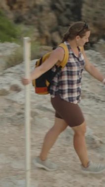 Dikey video. Denizin üzerindeki engebeli bir yamaç yolunda yürüyen bir kadın. Dalgaların yavaşça kayalara çarpmasıyla Alttaki Derin Mavi Su 'nun görüntüsünü alır..