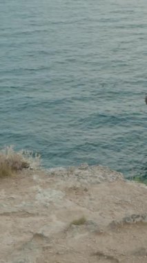 Dikey video. Okyanusun üzerindeki Rocky Patikası boyunca yürüyen bir yürüyüşçü. Uçurumun kenarındaki yol aşağıdaki suyun dramatik bir görüntüsünü sunuyor. Dik kayalar denize doğru iniyor..