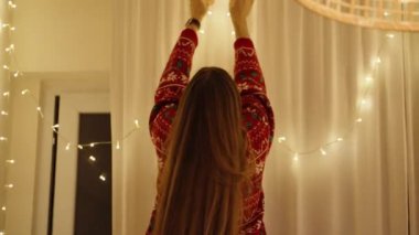 Kırmızı Şenlikli Süveter Giyen Bir Kadın Perdedeki İp Işıklarını Dikkatlice Ayarlıyor, Evinde Sıcak Tatil Ambiansı Oluşturuyor.