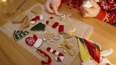 Kırmızı kazaklı bir kadın, Tahta bir tahtanın üzerinde süslü Noel kurabiyeleriyle çevrili zencefilli kurabiyeye beyaz buzlu şeker ekler..