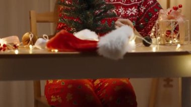 Kırmızı Noel Süveteri Giyen Kadın Süslü Işıkları Dekoratif bir kutunun üzerine diziyor Küçük bir Noel Ağacının yanına.