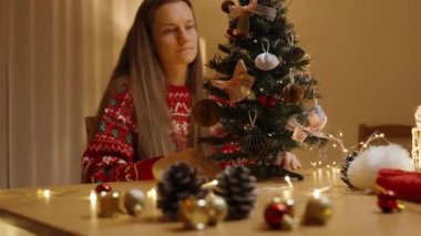 Kırmızı kazaklı bir kadın Noel ağacını dikkatlice inceliyor ve her şeyin mükemmel olmasını sağlıyor..