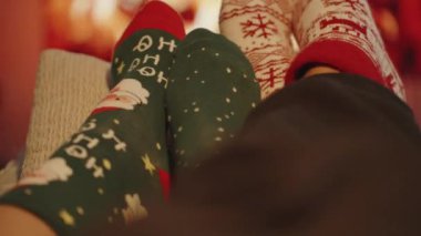 Bir erkek ve bir kadın şöminenin önünde dinleniyor, Ayakları Şenlikli Noel Çoraplarında.