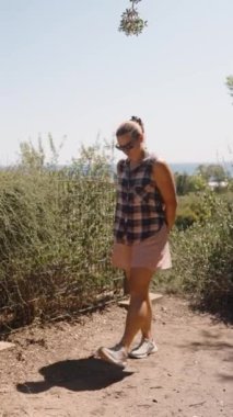 Dikey video. Bushy Fence 'in yanındaki Kirli Yol' da yürüyen bir kadın, Parlak Gök 'ün altındaki okyanusa bakıyor..