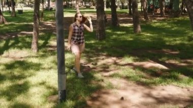 Ağaçlarla çevrili, güneş gözlüğü ve ekose gömlek takan bir kadın, güvenli bir şekilde gölgeli bir orman yolunda yürüyor..